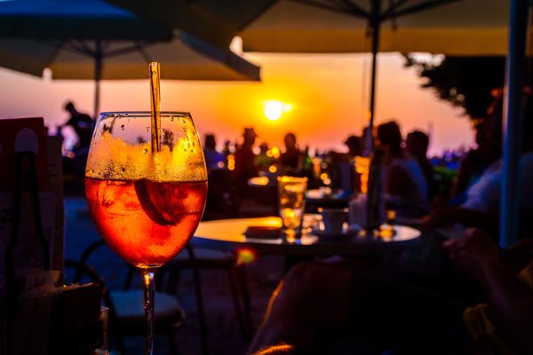 A glass of cold orange cocktail at the sunset on the table of a beach bar at the sunset, with blurry people arround having refreshements or partying on a summer evening, with copy space for text